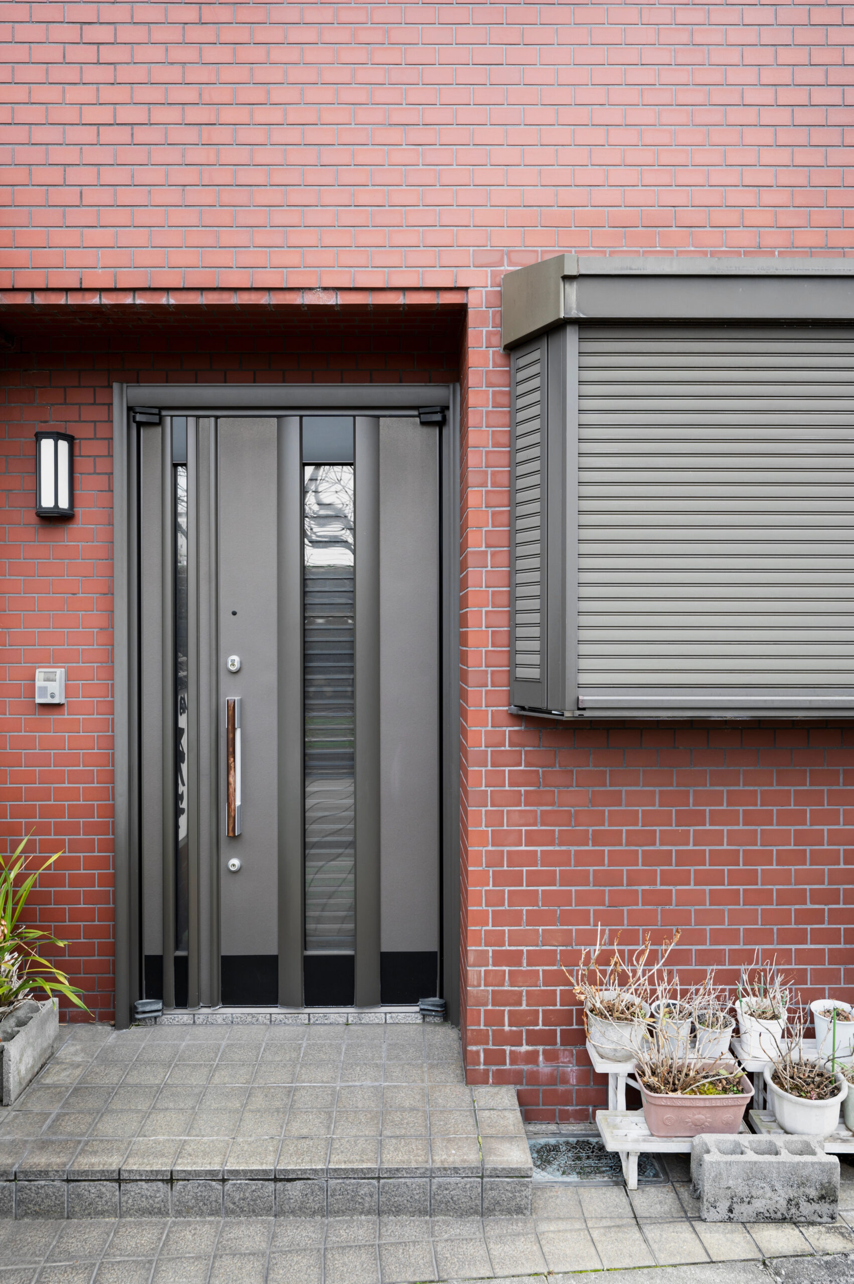 Sleek modern door with vertical glass panels on a red brick wall. A nearby shuttered window and potted plants add urban charm.