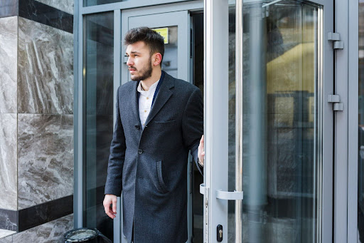 Man in a dark gray coat opening a modern glass door of a building.