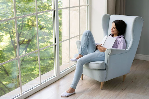A woman relaxes in a light-blue armchair, engrossed in a book near a large window, enjoying the view of the outdoors